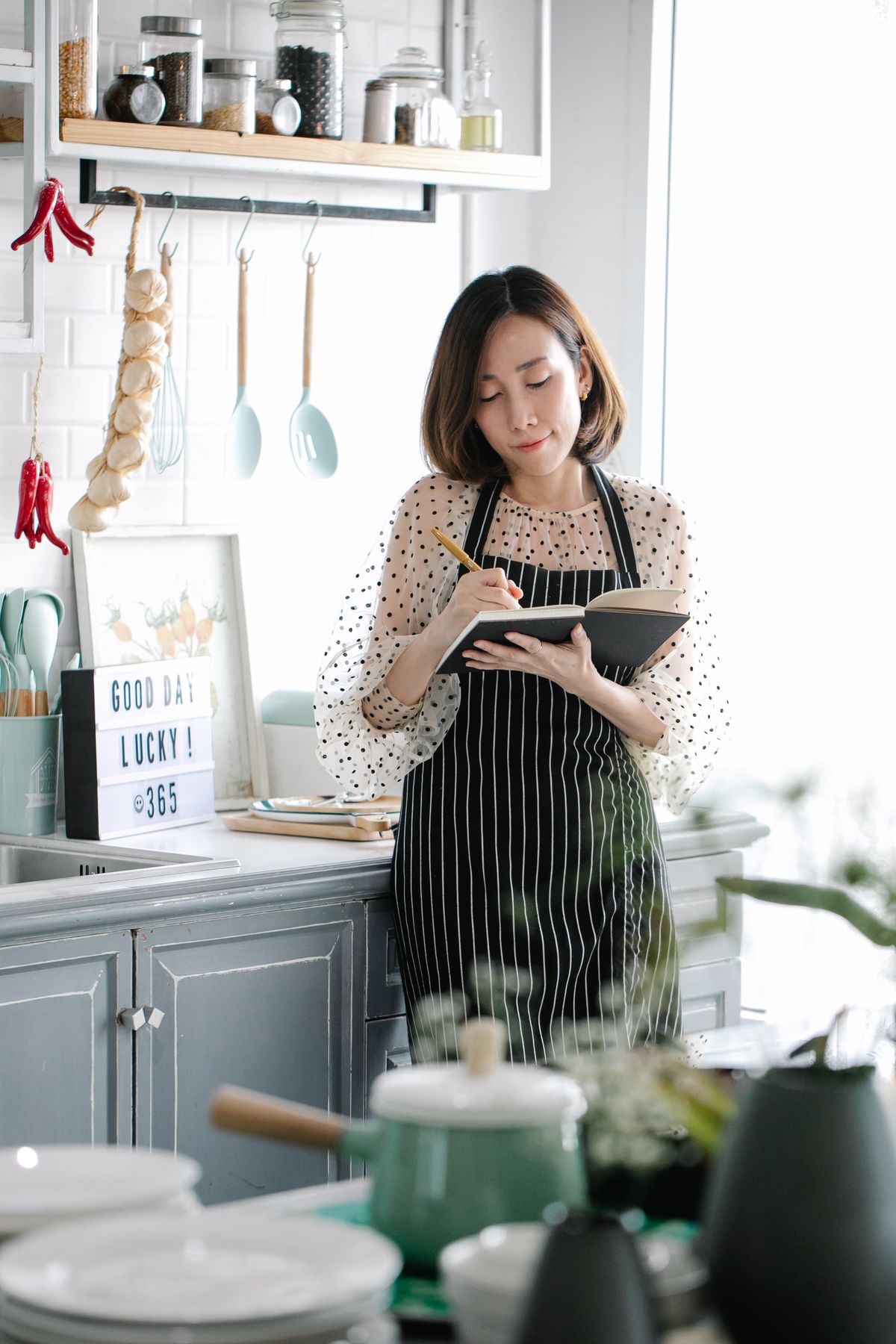 Woman in an apron writing Christmas hosting timeline notes in a journal at a kitchen counter with cooking utensils