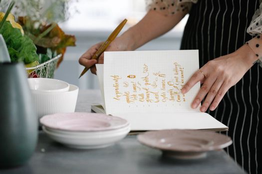 Woman writing a recipe in a notebook in a kitchen setting, surrounded by kitchenware.