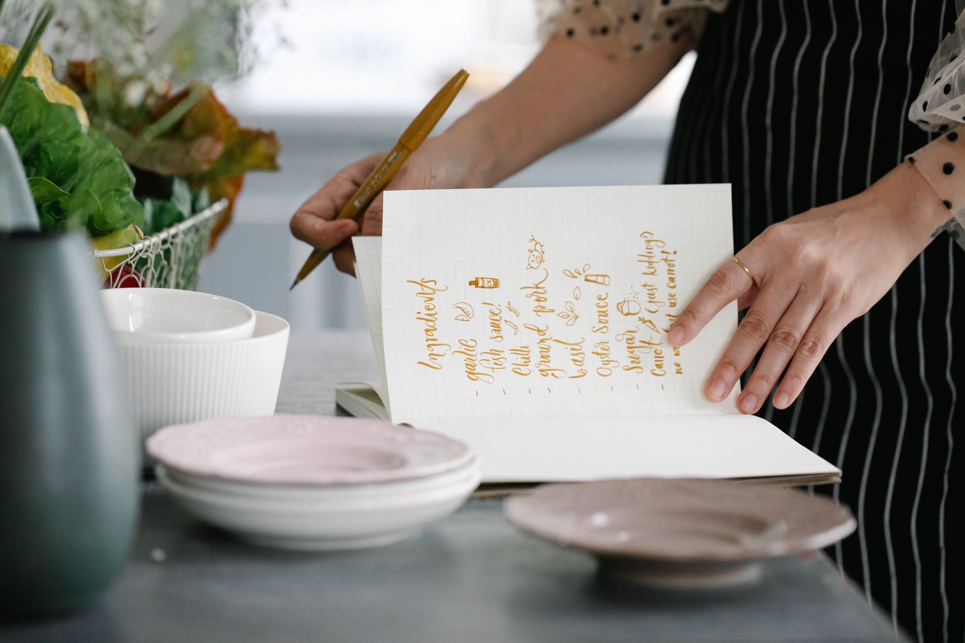 Woman writing a recipe in a notebook in a kitchen setting, surrounded by kitchenware.