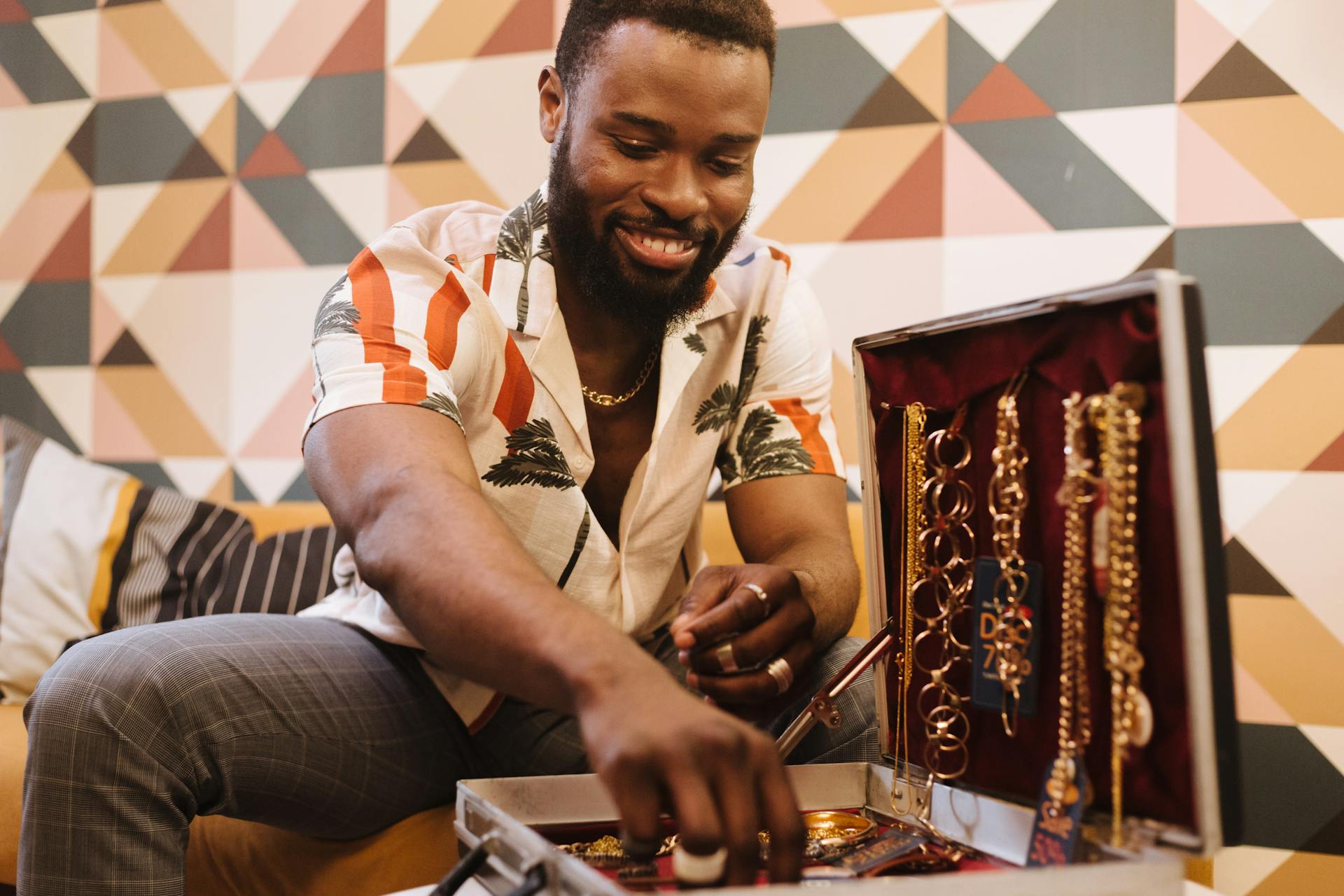 A cheerful man in trendy attire displays jewelry from an open case in an indoor setting.