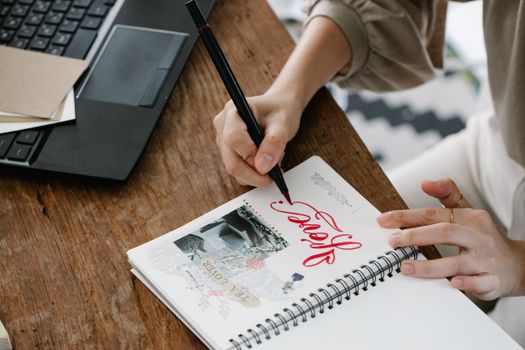 Close-up of a woman writing with red ink in a notebook on a wooden desk with a laptop.