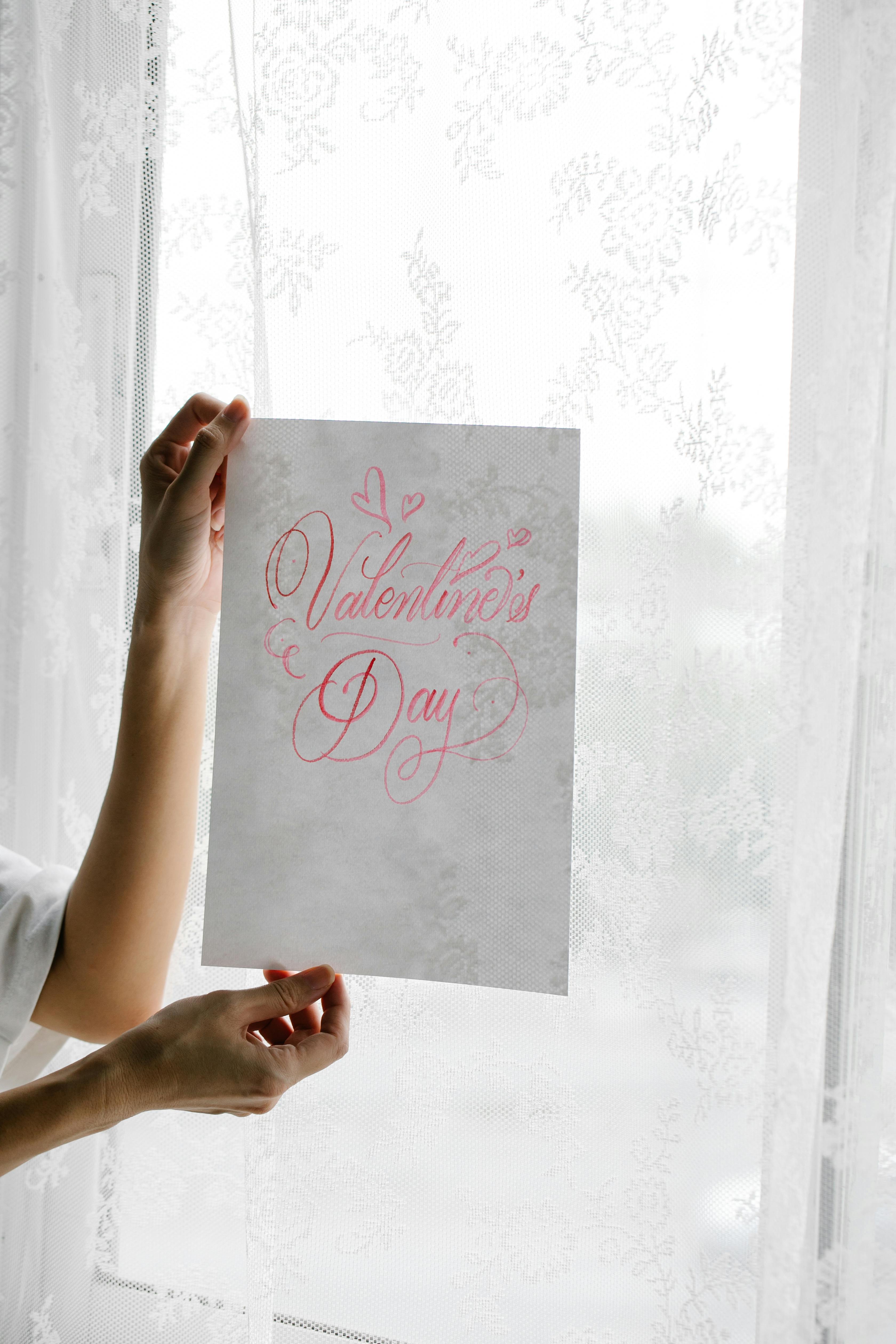 A person delicately holds a sheet with Valentine's Day hand lettering against a lace curtain backdrop.