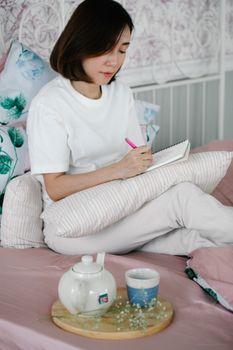 Woman sitting on a bed journaling with tea set nearby, creating a cozy, calm atmosphere.