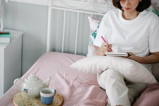 A woman in a white t-shirt takes notes in a notebook while sitting on a bed with tea beside her.
