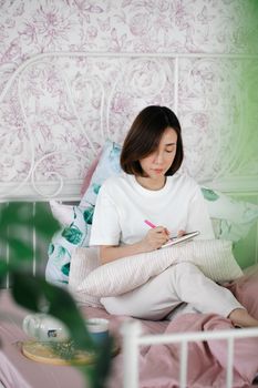 Woman in a white t-shirt writing in a notebook on a bed with floral wallpaper and pillows.