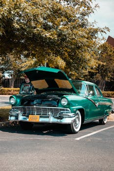 Vintage green car with open hood parked under trees. Perfect for classic car enthusiasts.