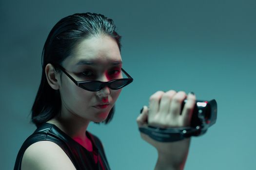 Portrait of a chic Asian woman wearing sunglasses and filming with a vintage camcorder under moody lighting.