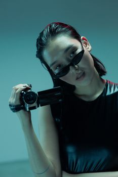 Stylish young Asian woman with sunglasses holding camcorder in a studio environment.