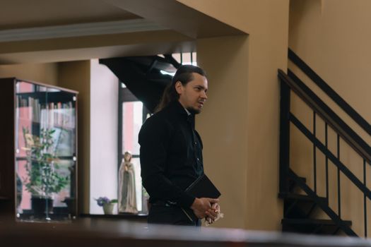 A priest in black attire holds a Bible inside a church, symbolizing faith and spirituality.