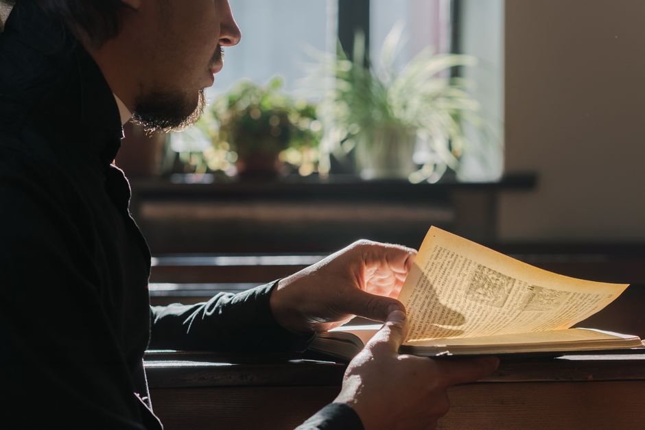 A man in black reads in a church, illuminated by window light, evoking spirituality and tranquility.