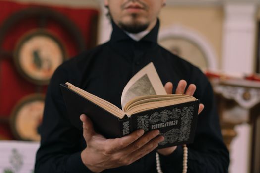 A religious figure in a black robe reading from a decorated sacred book indoors.