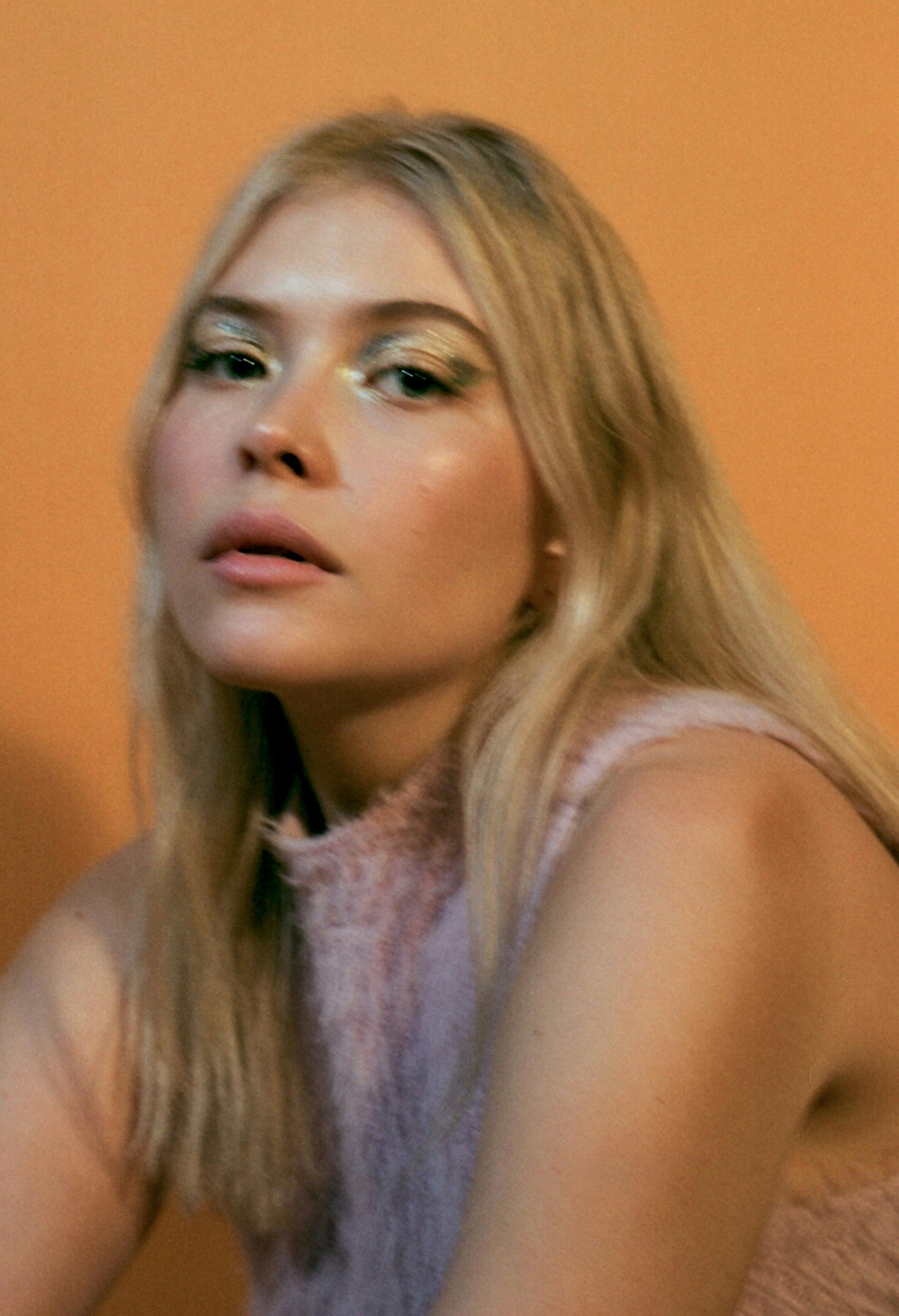 Blonde woman in a light pink outfit posing against a warm orange background, conveying a modern and artistic vibe.