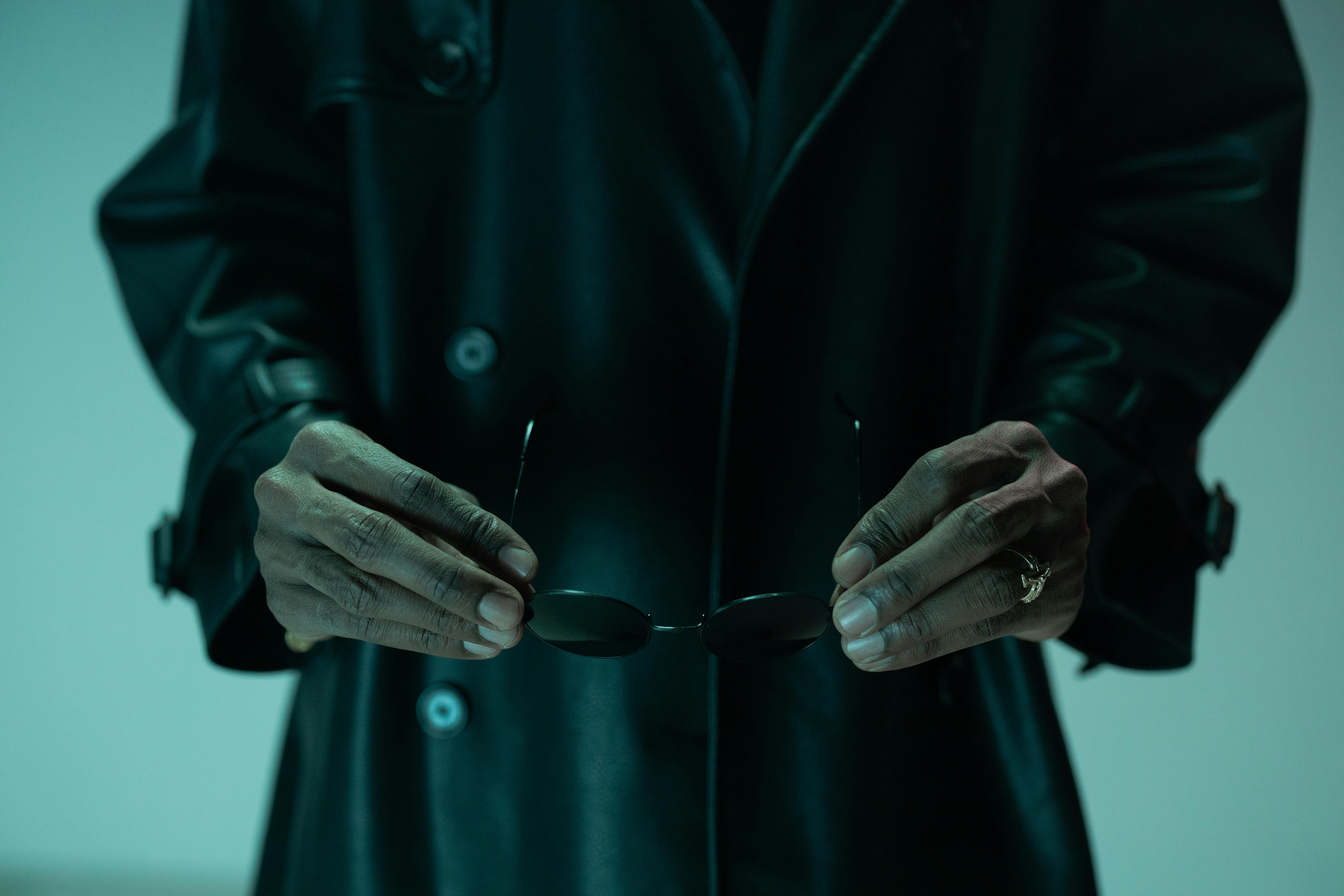 A mysterious close-up image of hands holding sunglasses, cloaked in a dark jacket under moody lighting.