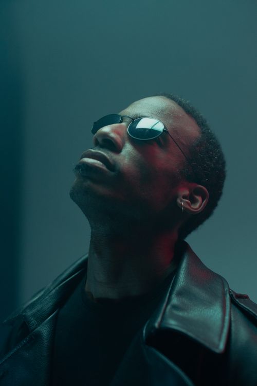 Free Portrait of a stylish man in a leather jacket and sunglasses gazing upward against a teal backdrop. Stock Photo