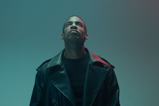 A man in a black leather jacket looks upwards against a teal background.