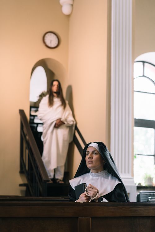 Free A nun praying in a chapel with another figure on the staircase behind her. Stock Photo