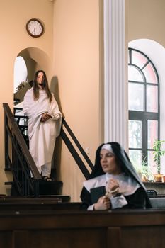 A nun praying in church while a woman descends the staircase, symbolizing devotion and faith.