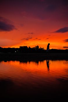 Silhouette of a person walking by a calm lake during a vibrant orange sunset.
