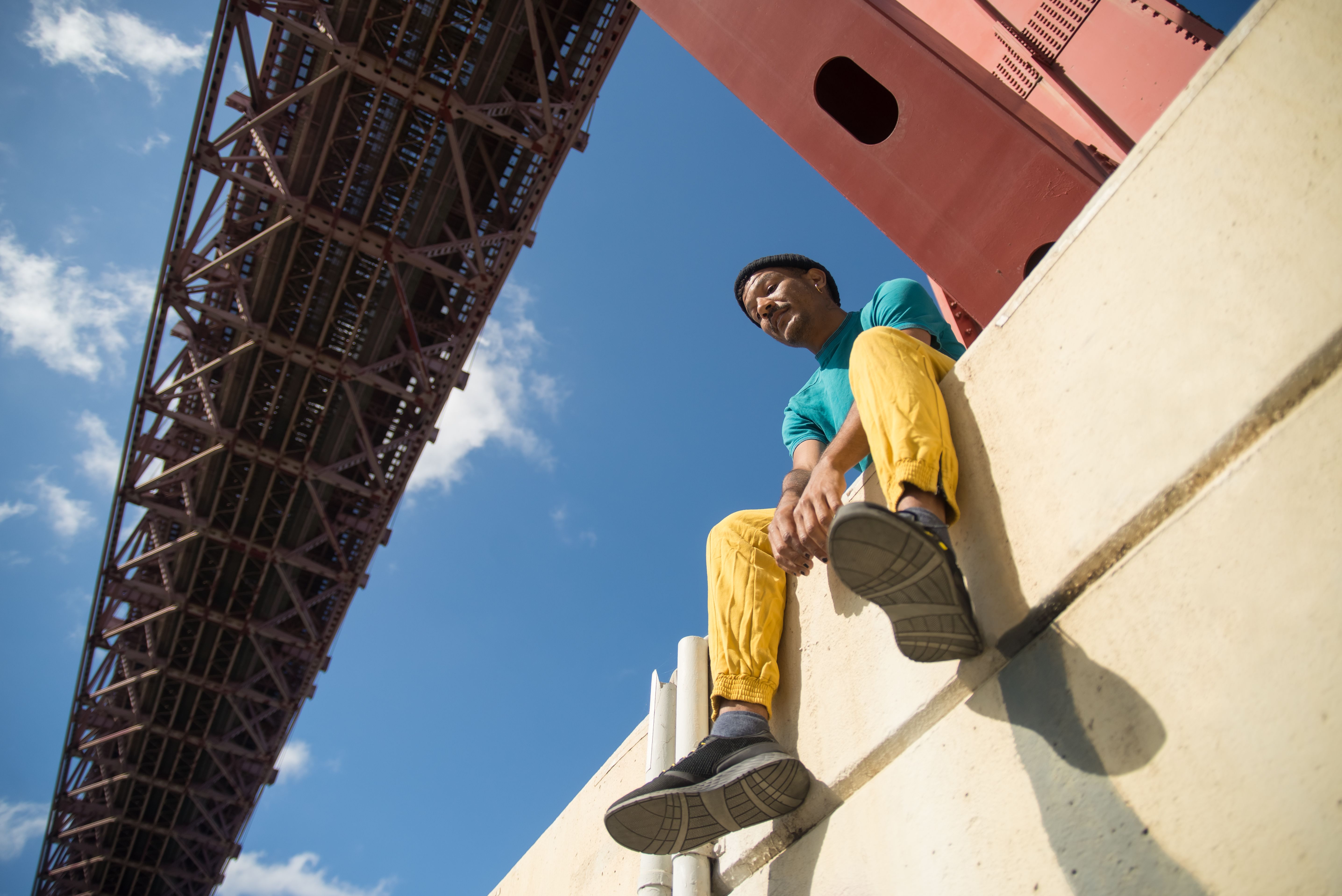 Man in colorful streetwear sitting under iconic bridge in Portugal, clear blue sky above.