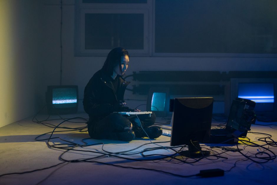 A focused woman sits in a dimly lit room surrounded by monitors and cables, creating a tech-savvy atmosphere.
