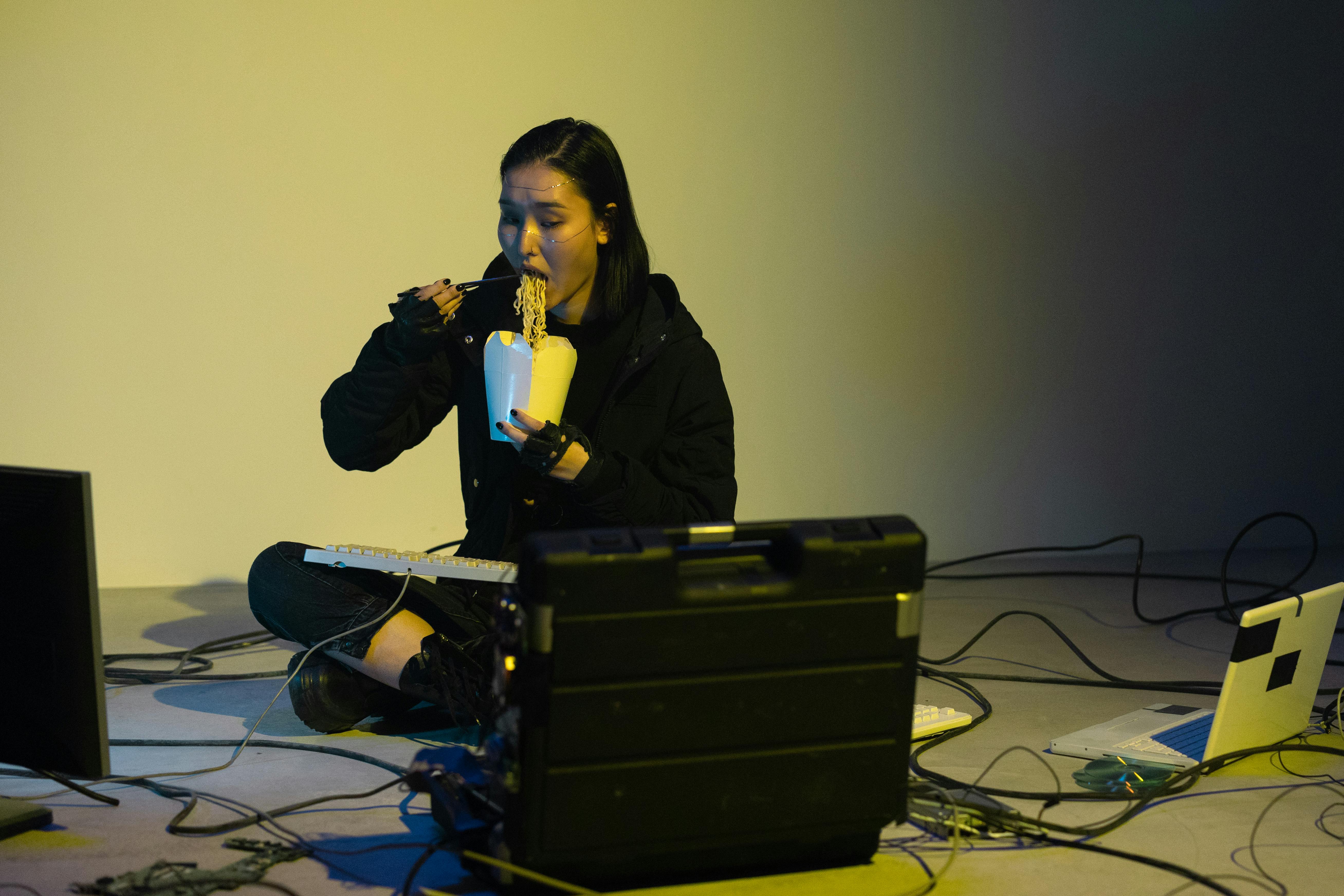 A young Asian woman multitasks by eating noodles while working on a laptop indoors.