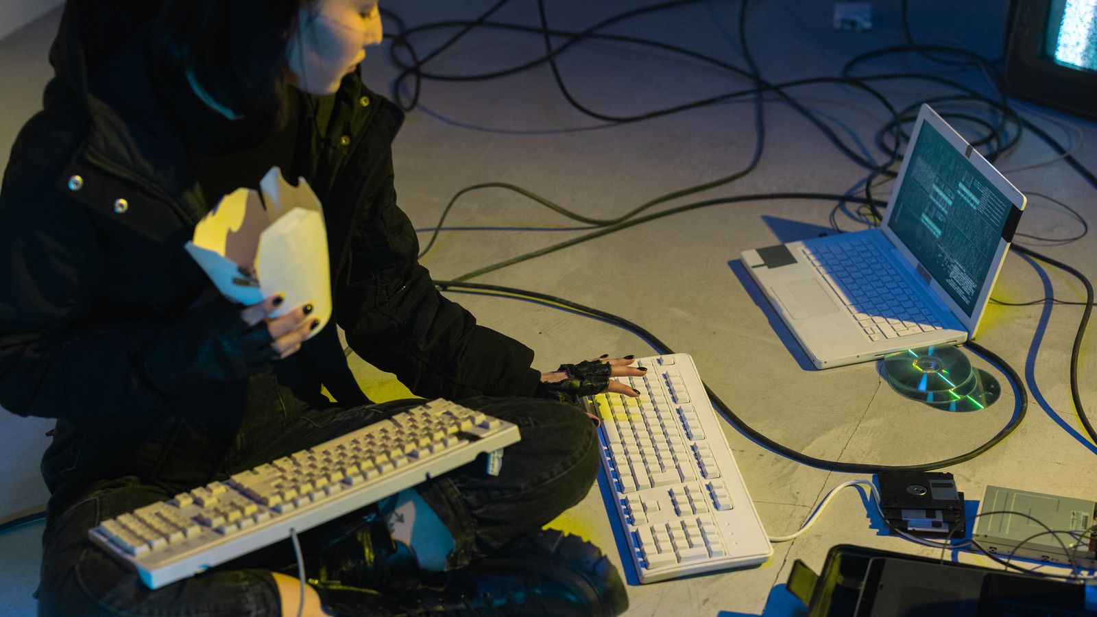 A young woman in a dark room surrounded by computers and cables, eating and typing on keyboards.