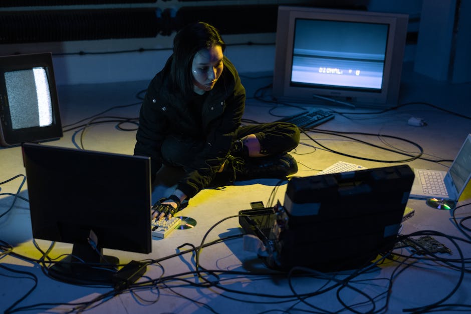 Asian woman working on multiple devices in a tech-heavy environment, illuminated by screen light.