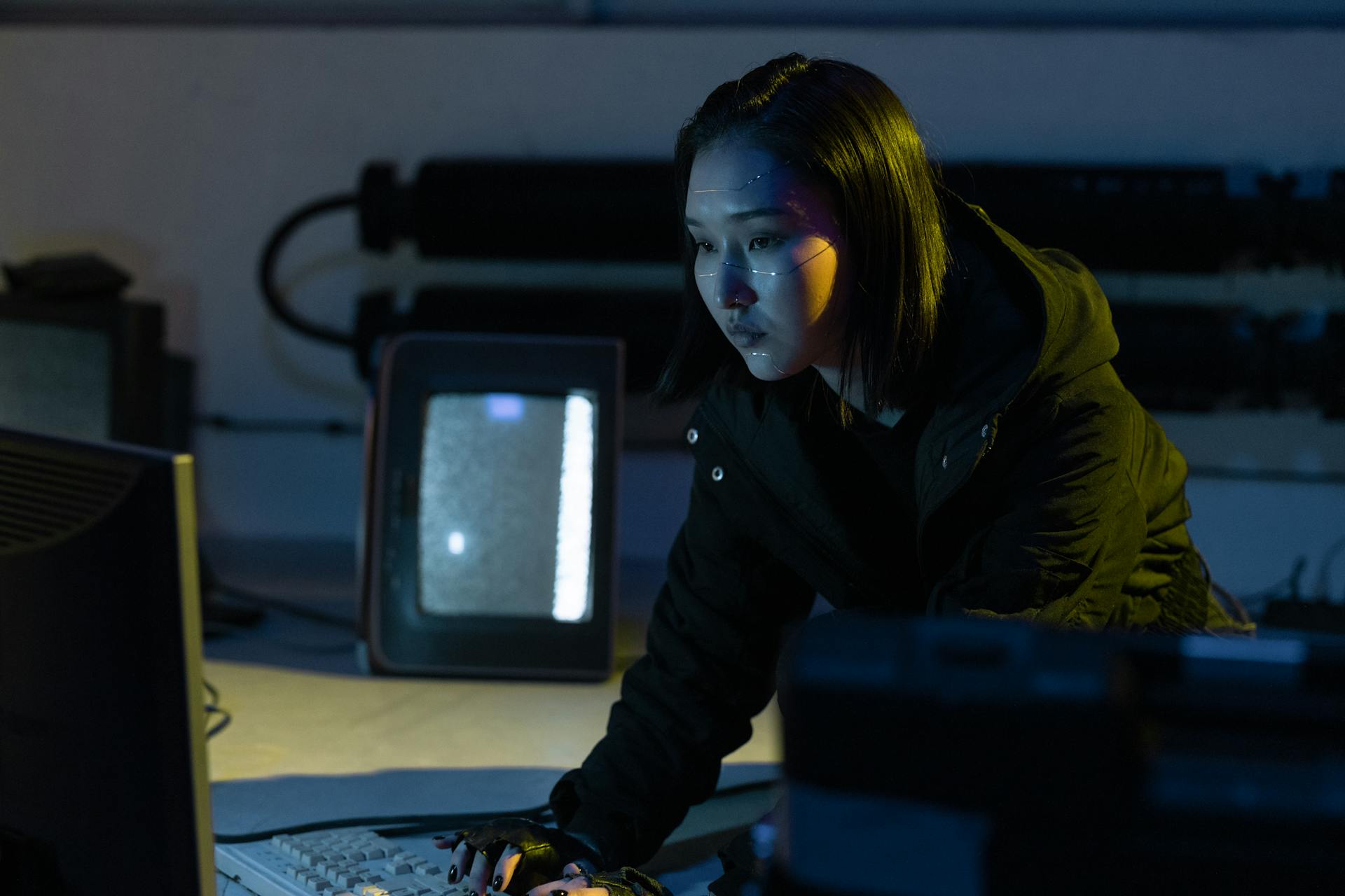 A woman working on a keyboard in a dimly lit room with tech equipment.