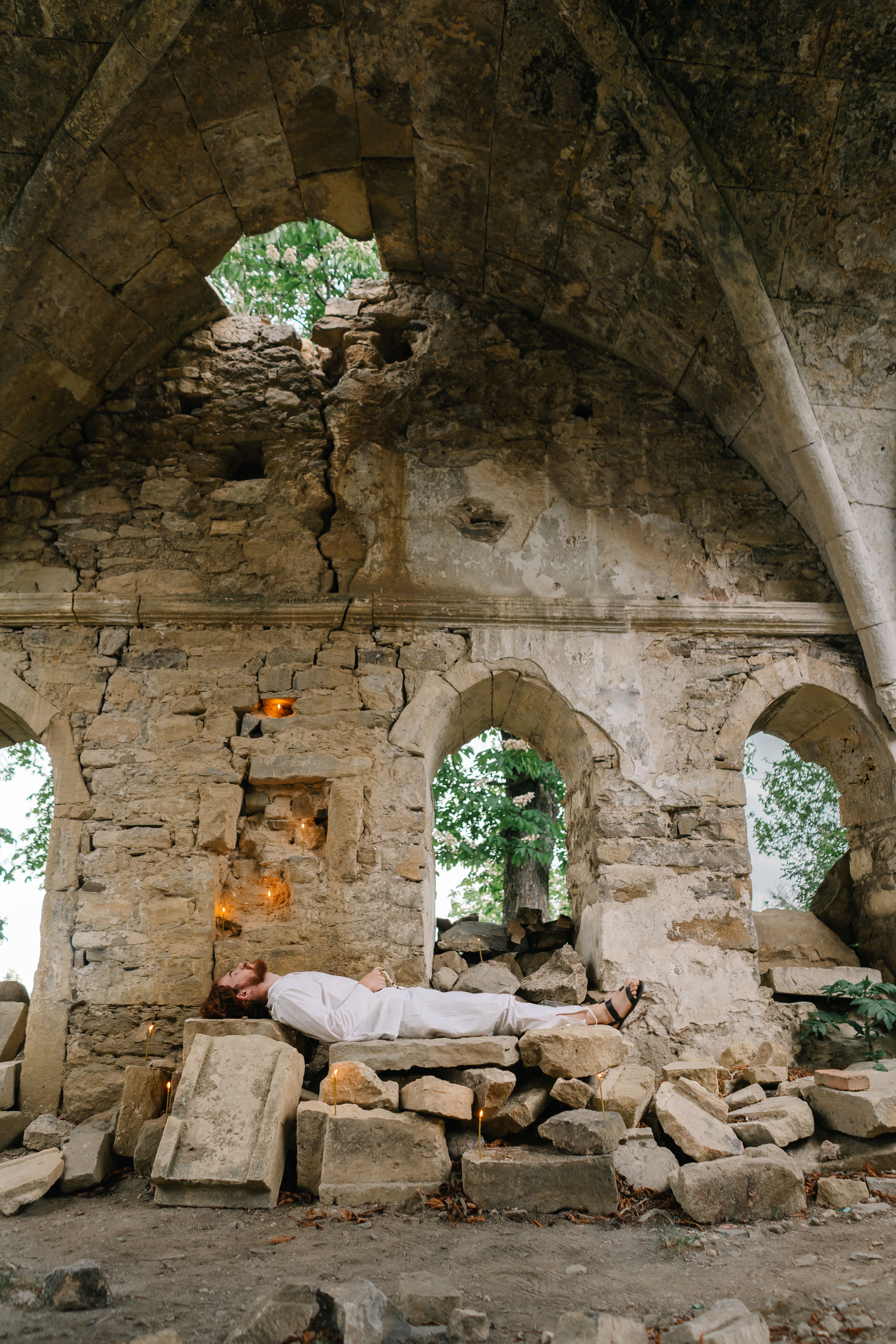 A serene scene of a man resting among ruins, surrounded by candles in a historic church setting.