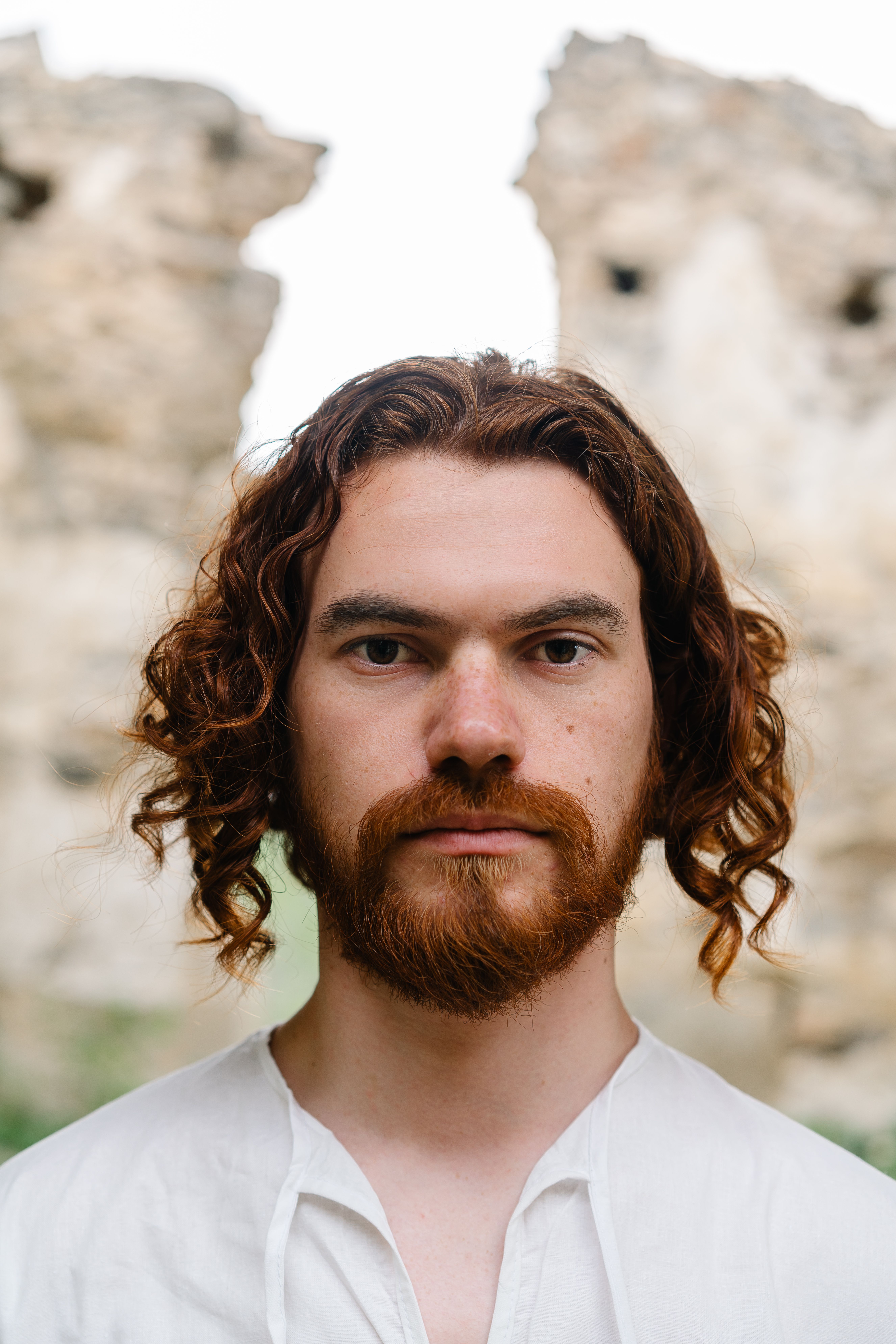 Portrait of a bearded man with curly hair against a rocky outdoor backdrop, conveying a natural and serene vibe.