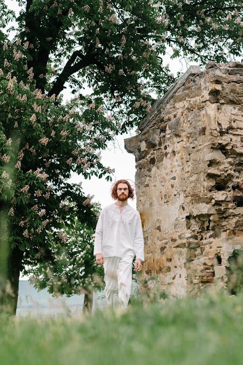 Man in White Clothes Walking on a Meadow near a Building Ruin 