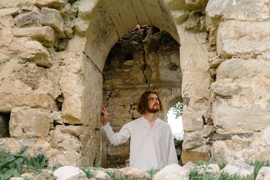 A man in a white shirt stands among ancient stone ruins, gazing thoughtfully.