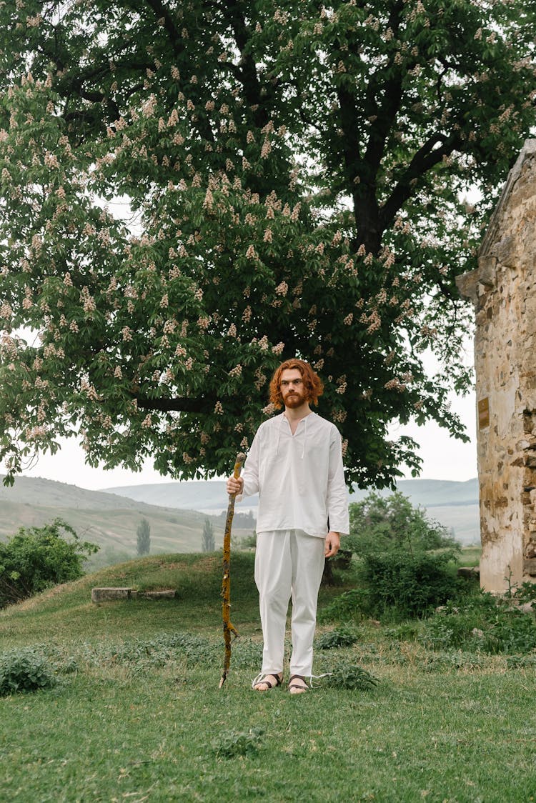 
A Bearded Man Wearing A White Long Sleeved Shirt And White Pants Holding A Stick