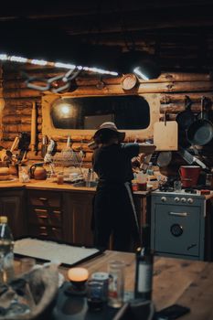 A woman in a hat cooking in a rustic kitchen with warm lighting and wooden decor.