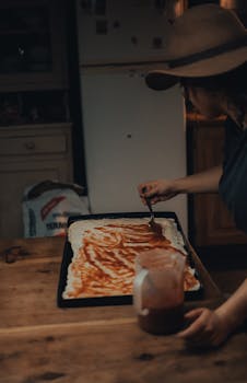 A woman spreads homemade tomato sauce on pizza dough in a cozy kitchen.