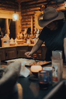 A woman in a rustic kitchen prepares pizza by candlelight, adding sauce to dough.