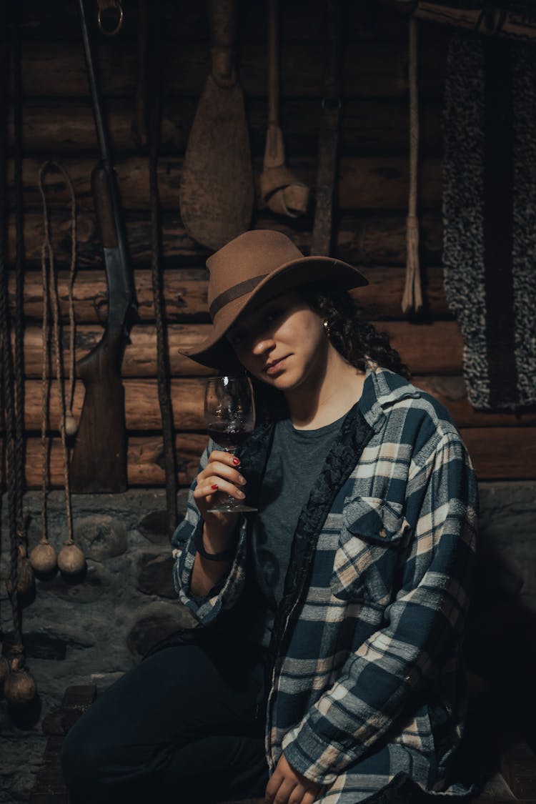 
A Woman In A Plaid Shirt And A Hat Holding A Glass Of Wine