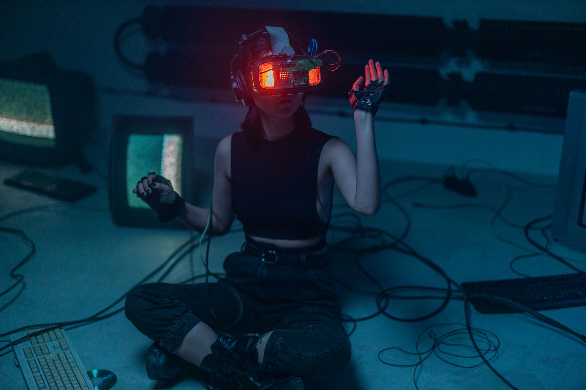 A woman wearing a VR headset sits on the floor surrounded by retro tech in a dark room.