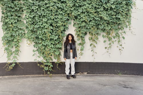Woman in Black Leather Jacket and White Pants Standing Beside Wall With Plants 