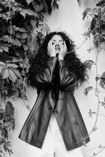 Monochrome portrait of a stylish woman in a leather jacket and curly hair, against a leafy vine wall.