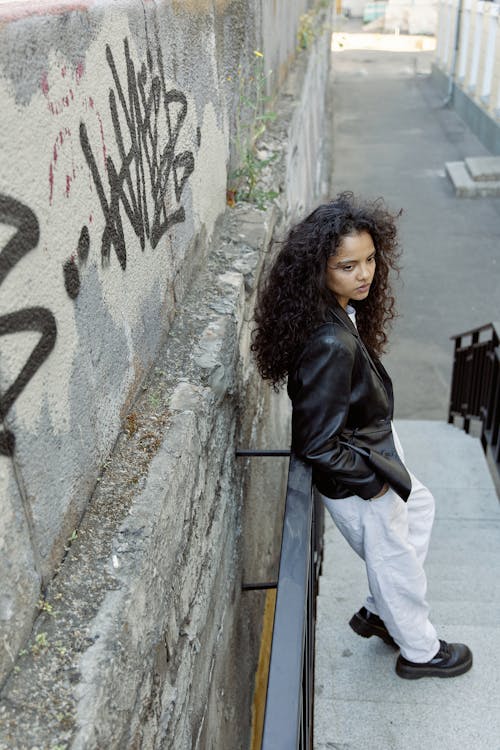 A Woman in Black Leather Jacket and White Pants Standing Beside Concrete Wall
