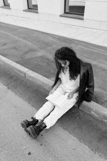 Black and white photo of a woman sitting on the curb reading a book. Urban scene.