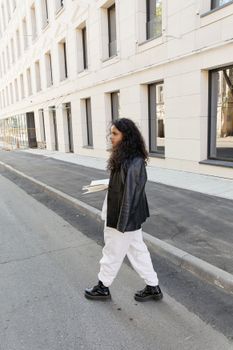 Fashionable woman with curly hair walking on an urban street in a black leather jacket and white pants.