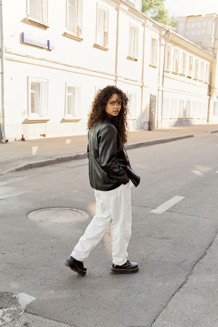 A young woman in a leather jacket walks on a sunny city street, showcasing urban fashion.