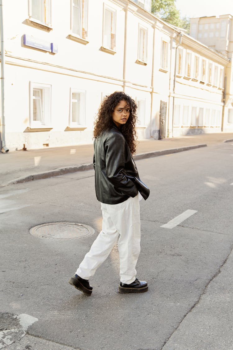 Young Woman In Black Leather Jacket And White Pants Walking On Street