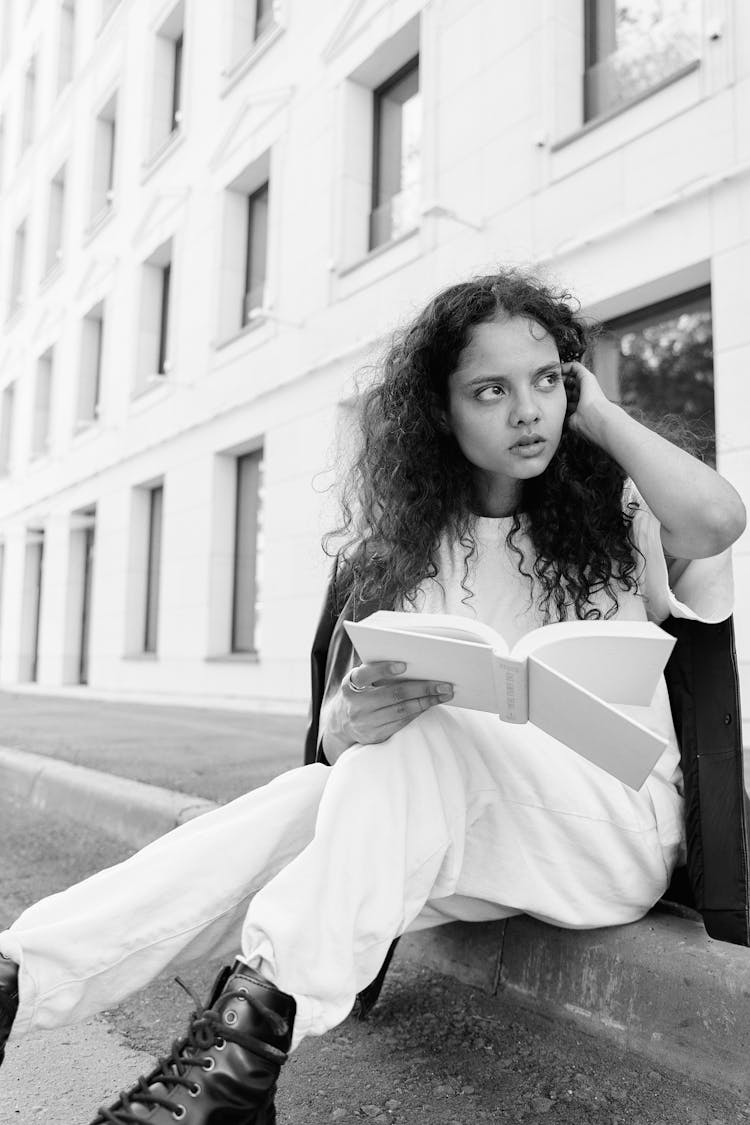 Grayscale Photo Of Woman Sitting On Gutter While Holding A Book