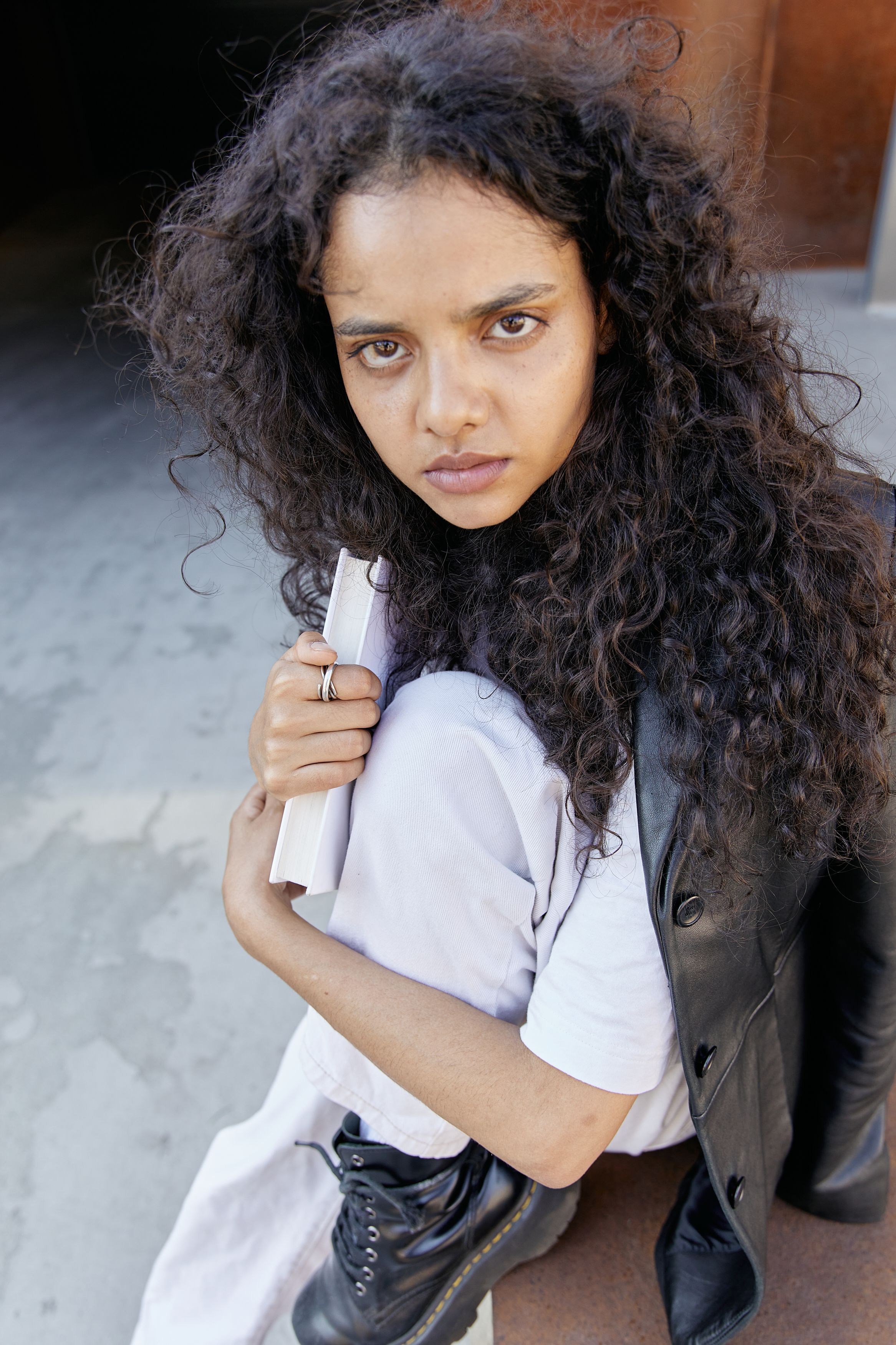 Portrait of a young woman with curly hair holding a book, exuding a fashionable and introspective vibe.