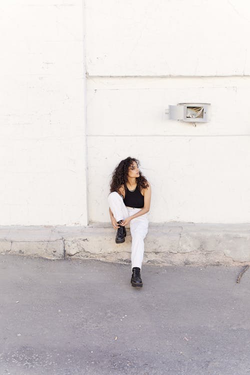 Woman in Black Tank Top and White Pants Sitting on Concrete Floor