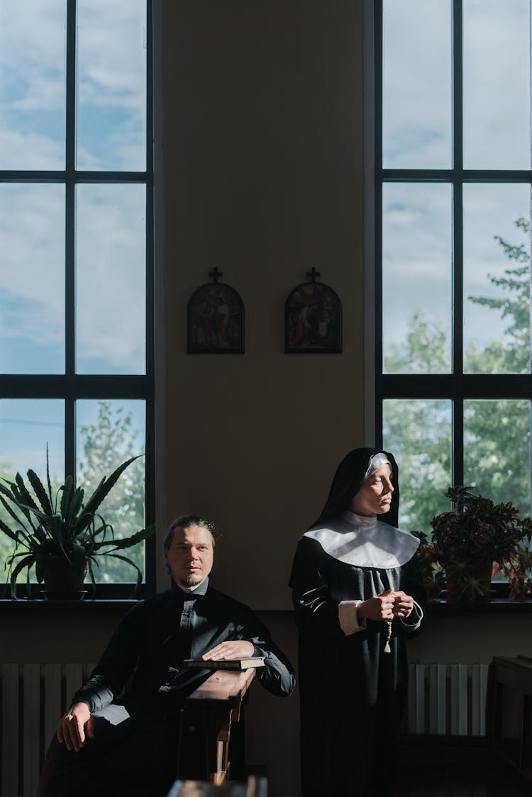 Nun Standing And Holding Prayer Beads Beside A Priest Sitting On Church Pew