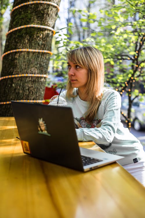 Blond Woman Sitting at a Wooden Table with Laptop and Trees in Background